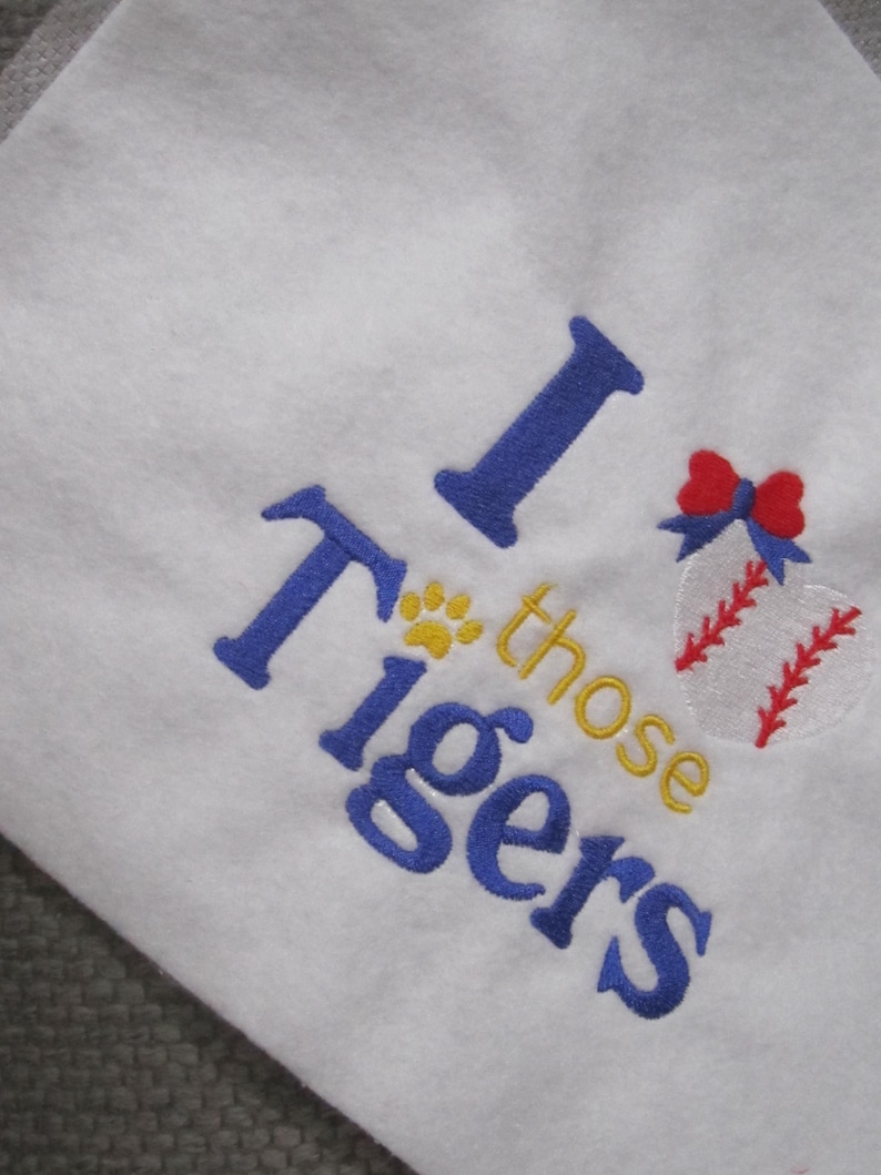 May include: White fabric with blue embroidered text that reads "I those Tigers". A baseball with red stitching and a red bow is embroidered on the right side. A yellow paw print is above the word "those".