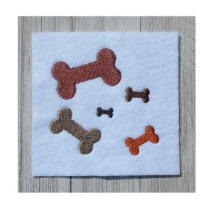 May include: A white felt patch with four embroidered dog bones in different sizes and colors: orange, brown, and tan.