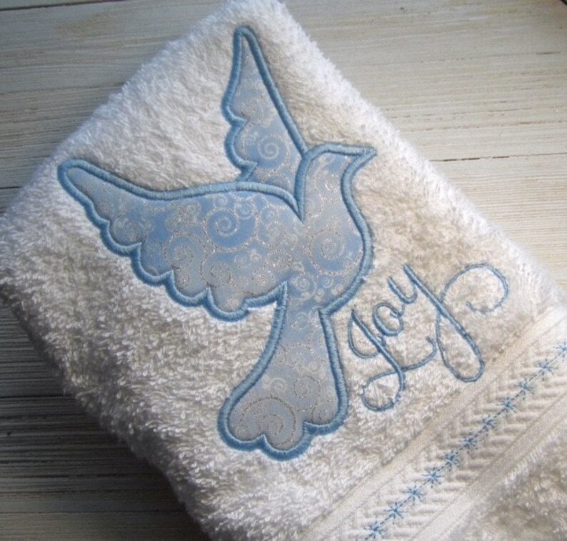 May include: White bath towel with a blue embroidered dove and the word "Joy" in cursive script.