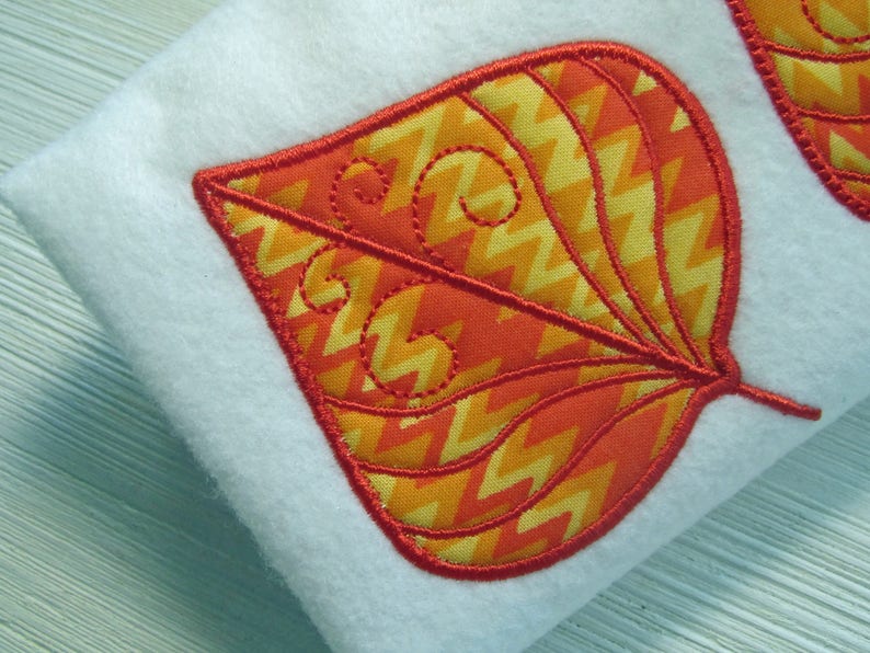 K&ouml;nnte beinhalten: Nahaufnahme eines blattf&ouml;rmigen Applikationsmusters in leuchtenden Herbstfarben. Das Blatt hat ein Zickzackmuster in Orange und Gelb, umrandet von Rot. Die Applikation befindet sich auf einem wei&szlig;en Hintergrund.