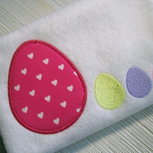 Puede incluir: Una tela blanca con huevos bordados en rosa, verde, morado y rosa. El huevo rosa tiene un patrón de corazones blancos.