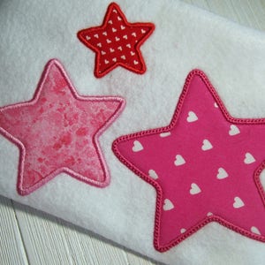 May include: Three embroidered stars in pink and red with white heart patterns. The stars are sewn onto a white fabric background.