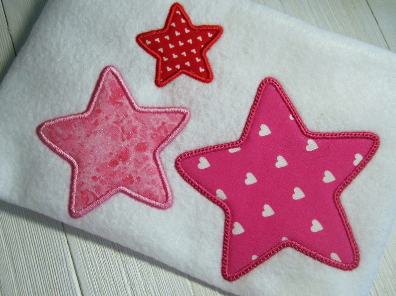 May include: Three embroidered stars in pink and red with white heart patterns. The stars are sewn onto a white fabric background.