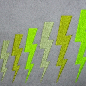 May include: A gray background with a pattern of embroidered lightning bolts in varying shades of green. The bolts increase in size from left to right, creating a visual gradient. The embroidery has a textured appearance.