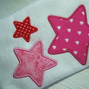 May include: Three embroidered stars on a white fabric background. The largest star is pink with white polka dots, the medium star is pink with a glittery texture, and the smallest star is red with white polka dots.