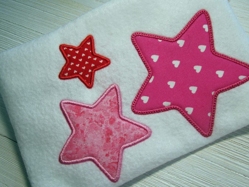 May include: Three embroidered stars on a white fabric background. The largest star is pink with white polka dots, the medium star is pink with a glittery texture, and the smallest star is red with white polka dots.