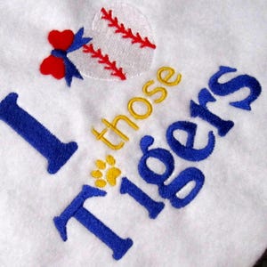 May include: White fabric with embroidered text and baseball-themed design. The text reads "I those Tigers" in blue and gold thread. A baseball with red stitching and a red bow are also embroidered.