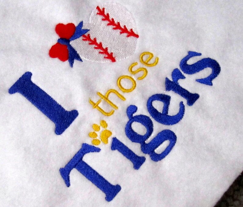 May include: White fabric with embroidered text and baseball-themed design. The text reads "I those Tigers" in blue and gold thread. A baseball with red stitching and a red bow are also embroidered.