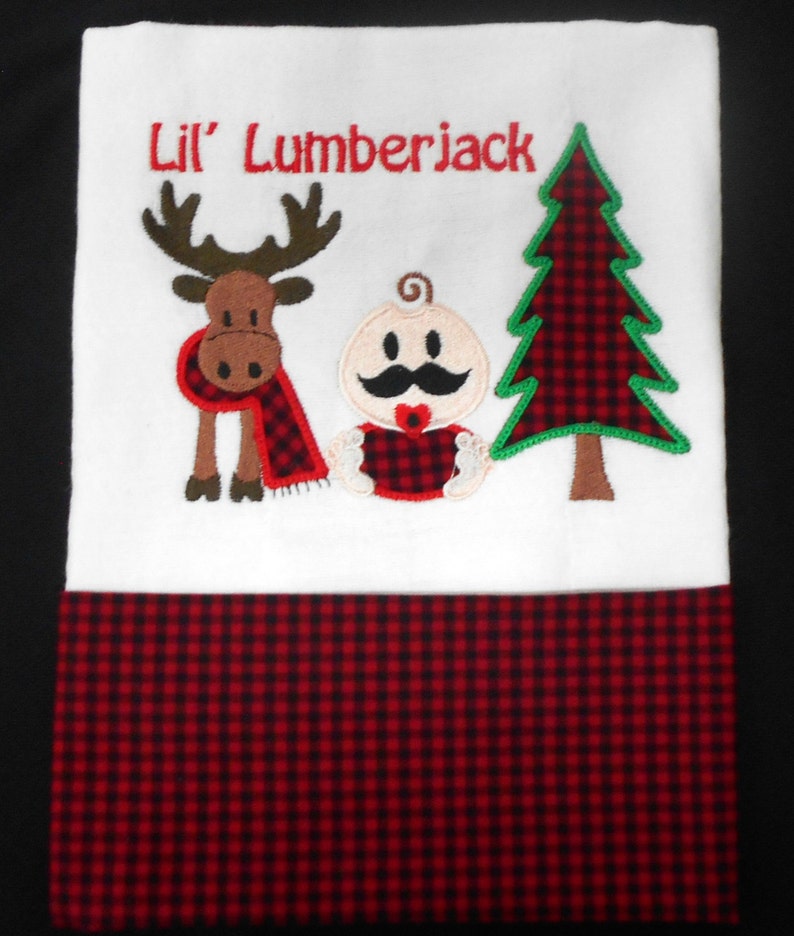 May include: White baby burp cloth with red and black buffalo plaid trim. The cloth features an embroidered design of a baby with a mustache, a moose, and a Christmas tree. The text "Lil' Lumberjack" is embroidered above the design.