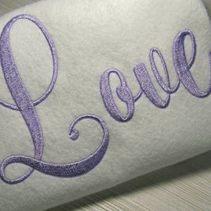 Può includere: Un cuscino bianco con la parola "Love" ricamata in un carattere corsivo di colore lilla chiaro.