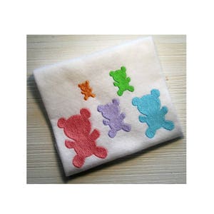 Pode incluir: Um tecido branco com cinco silhuetas de ursos de pelúcia bordadas em diferentes cores: laranja, verde, roxo, rosa e azul.