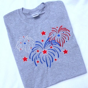 May include: A gray t-shirt with an embroidered design of red, white, and blue fireworks. The fireworks are surrounded by red stars.