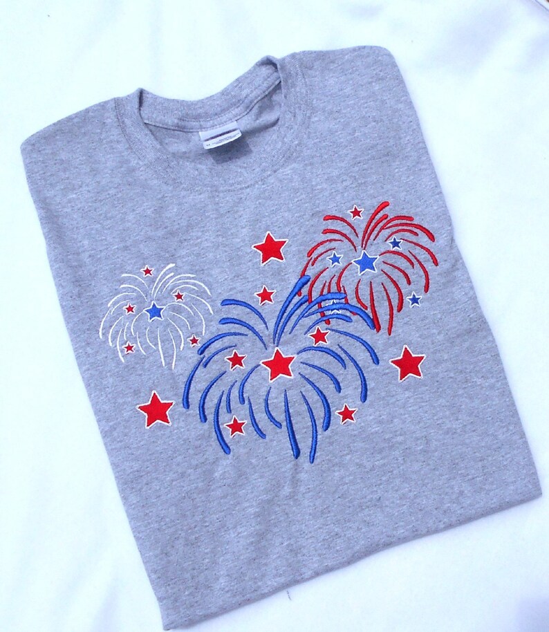 May include: A gray t-shirt with an embroidered design of red, white, and blue fireworks. The fireworks are surrounded by red stars.