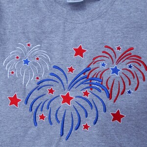 May include: A gray t-shirt with an embroidered design of three red, white, and blue fireworks. The fireworks are surrounded by smaller red stars.