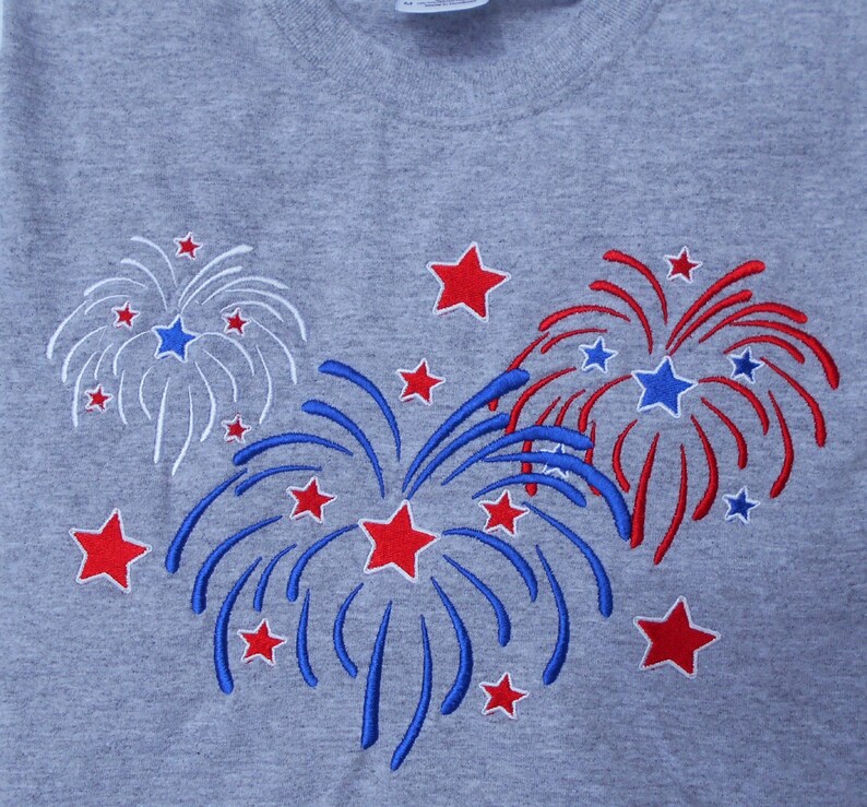 May include: A gray t-shirt with an embroidered design of three red, white, and blue fireworks. The fireworks are surrounded by smaller red stars.