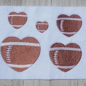 K&ouml;nnte beinhalten: F&uuml;nf braune Filz-Herz-Patches mit wei&szlig;en N&auml;hten, die an Fu&szlig;b&auml;lle erinnern. Die Patches sind in einem Halbkreis auf wei&szlig;em Hintergrund angeordnet.