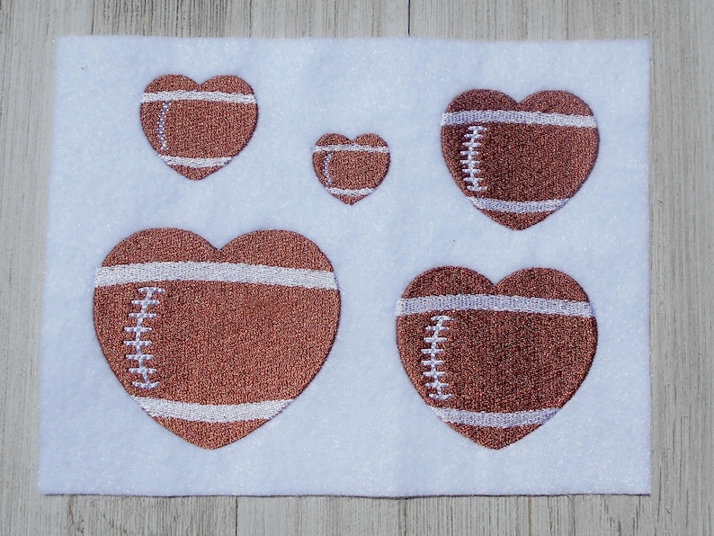 K&ouml;nnte beinhalten: F&uuml;nf braune Filz-Herz-Patches mit wei&szlig;en N&auml;hten, die an Fu&szlig;b&auml;lle erinnern. Die Patches sind in einem Halbkreis auf wei&szlig;em Hintergrund angeordnet.