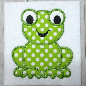 May include: Green felt applique of a frog with white polka dots and black eyes. The frog is sitting with its legs outstretched.
