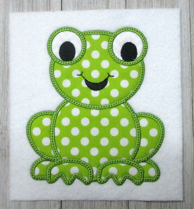 May include: Green felt applique of a frog with white polka dots and black eyes. The frog is sitting with its legs outstretched.