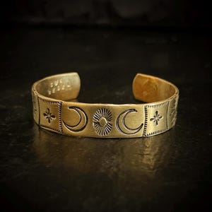 May include: A gold-colored cuff bracelet with celestial designs. The bracelet features embossed crescent moons, a sunburst, and star-like symbols. The open-ended design allows for easy wear. The bracelet is displayed on a dark surface.