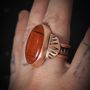 Puede incluir: Anillo de cobre con una gran piedra de jaspe rojo-marrón ovalada con vetas oscuras. El anillo tiene una banda decorativa con detalles grabados y un engaste texturizado. Una pieza de joyería llamativa.