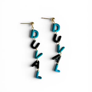 May include: A pair of dangling earrings spelling out "DUVAL" in a vertical arrangement. The letters are a combination of black and teal, with gold-colored earring posts. The earrings are set against a plain white background.
