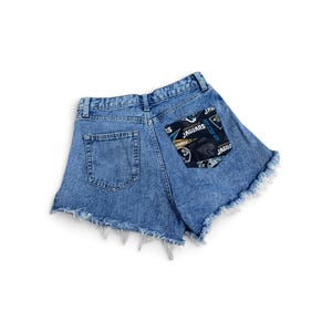 May include: Blue denim shorts with a frayed hem and a high-waisted cut. The back features two pockets, one of which is accented with a black fabric patch displaying the Jacksonville Jaguars logo, team name, and various pennant-style graphics.