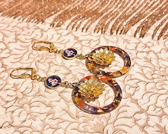 Könnte beinhalten: Goldfarbene Ohrringe mit floralem Design. Jeder Ohrring hat einen kreisförmigen Reifen mit farbenfrohem Muster, eine goldene Blume und einen kleinen runden Anhänger mit rosa Blumendesign. Die Ohrringe werden auf einer gemusterten Oberfläche präsentiert.