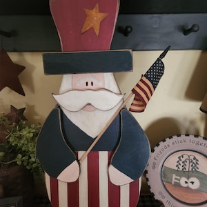 May include: A patriotic wooden figure of a man in a hat, holding a small American flag. The figure has a white mustache and is painted in red, white, blue, and tan. The hat has a gold star.