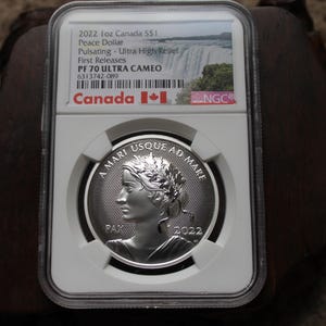 Peut inclure: Une pièce de monnaie canadienne Paix Dollar de 2022, encapsulée dans un étui de protection. La pièce présente un profil de femme avec une couronne de laurier et l'inscription "A MARI USQUE AD MARE". L'étui comprend des détails comme "PF 70 ULTRA CAMEO".