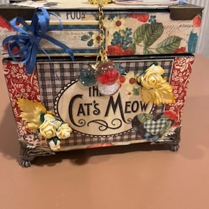 May include: A decorative box with a floral design and the words "The Cat's Meow" on the front. The box has a white knob and a gold chain with decorative beads.