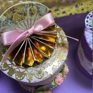 Op de afbeelding: Een kleine, ronde, decoratieve doos met een roze lint en een gouden waaier-vormige versiering. De doos is versierd met een wit kantpatroon en roze bloemen. De doos staat op een paarse ondergrond.