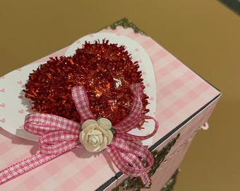 May include: A pink and white checkered box with a red glitter heart and a pink and white gingham ribbon. The box has a decorative metal clasp and feet.