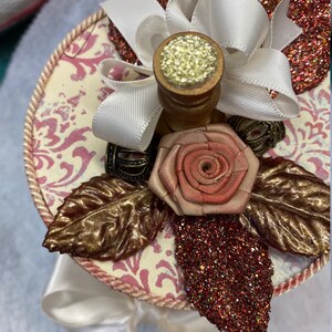 May include: A white and pink decorative box with a gold and brown leaf design, a pink rose, a white ribbon, and gold glitter accents.