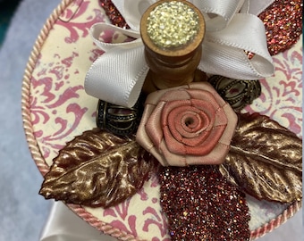 May include: A white and pink decorative box with a gold and brown leaf design, a pink rose, a white ribbon, and gold glitter accents.