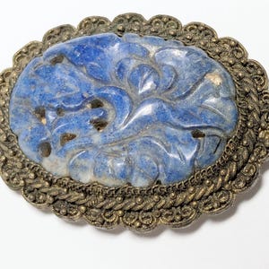 May include: Oval-shaped vintage brooch with a carved blue stone center featuring a floral design. The stone is set in a detailed, ornate silver-toned metal frame with a scalloped edge. A unique piece of jewelry.