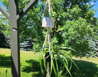 May include: A white macrame plant hanger with teal beads hangs from a wooden pergola. Two potted plants are suspended in the hanger. The plants are green and leafy.