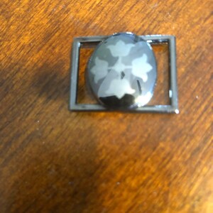 May include: A silver-toned metal charm with a black oval-shaped design featuring a stylized flower with four petals. The charm is framed by a square metal border.