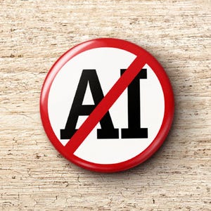 May include: A round button with a white background and a red border. The letters "AI" are printed in black with a red slash through them. The button is on a light brown, textured wooden surface.