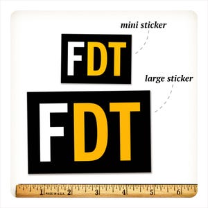 Może przedstawiać: Dwie czarne prostokątne naklejki z literami "FDT" w kolorze białym i ż&oacute;łtym. Mniejsza naklejka jest oznaczona jako "mini sticker", a większa jako "large sticker". Na dole znajduje się linijka w calach, około 15 cm.
