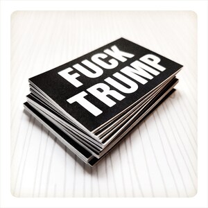 May include: A stack of black rectangular stickers with the words "FUCK TRUMP" printed in bold white capital letters. The stickers are stacked on a white surface with subtle vertical lines.