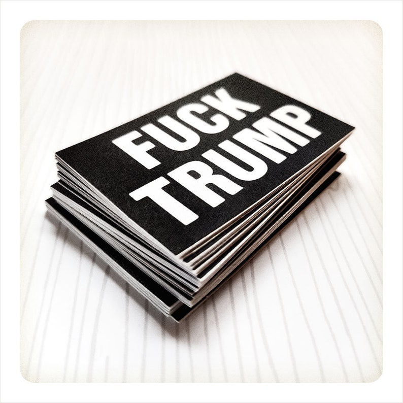 May include: A stack of black rectangular stickers with the words "FUCK TRUMP" printed in bold white capital letters. The stickers are stacked on a white surface with subtle vertical lines.