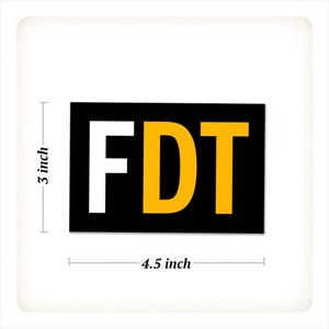 Może przedstawiać: Prostokątna naklejka z literami "FDT" pogrubioną czcionką. Litera "F" jest biała, "D" jest ż&oacute;łta, a "T" jest ż&oacute;łta. Tło jest czarne. Naklejka ma 11,4 cm szerokości i 7,6 cm wysokości.