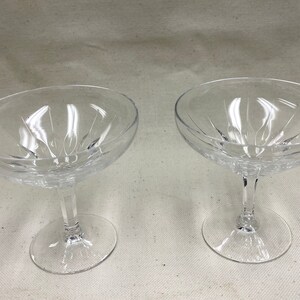 May include: Two clear glass champagne coupes with a delicate, faceted design. The coupes are empty and have a classic shape.