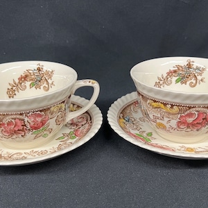 May include: Two matching teacups and saucers. The cups and saucers are cream-colored with a floral and paisley design in shades of pink, brown, and yellow. The cups have a curved handle and a textured rim. The saucers have a scalloped edge.