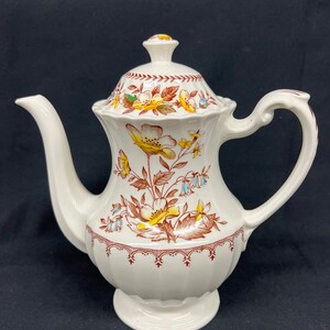 May include: A white ceramic teapot with a floral design. The teapot has a brown and yellow floral pattern with blue accents. The teapot has a lid and a handle.