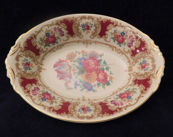 May include: An oval serving dish with a white background and a red and gold floral pattern. The dish has a scalloped edge and two handles.