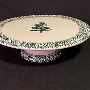 May include: A white ceramic cake stand with a green and red Christmas tree design. The rim is decorated with a green speckled pattern. The base is white with a matching green speckled pattern.
