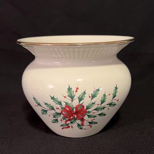 May include: Cream-colored ceramic vase with a gold rim and a festive holly and red bow design. The vase has a rounded body and a slightly flared top, perfect for holiday decor.