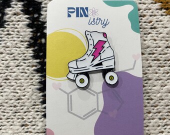 Könnte beinhalten: Weißer Rollschuh-Emaille-Pin mit einem pinkfarbenen Blitz-Design. Der Pin befindet sich auf einer weißen Karte mit dem Text "PINistry" und "Making Reactions Happen Since 2023".